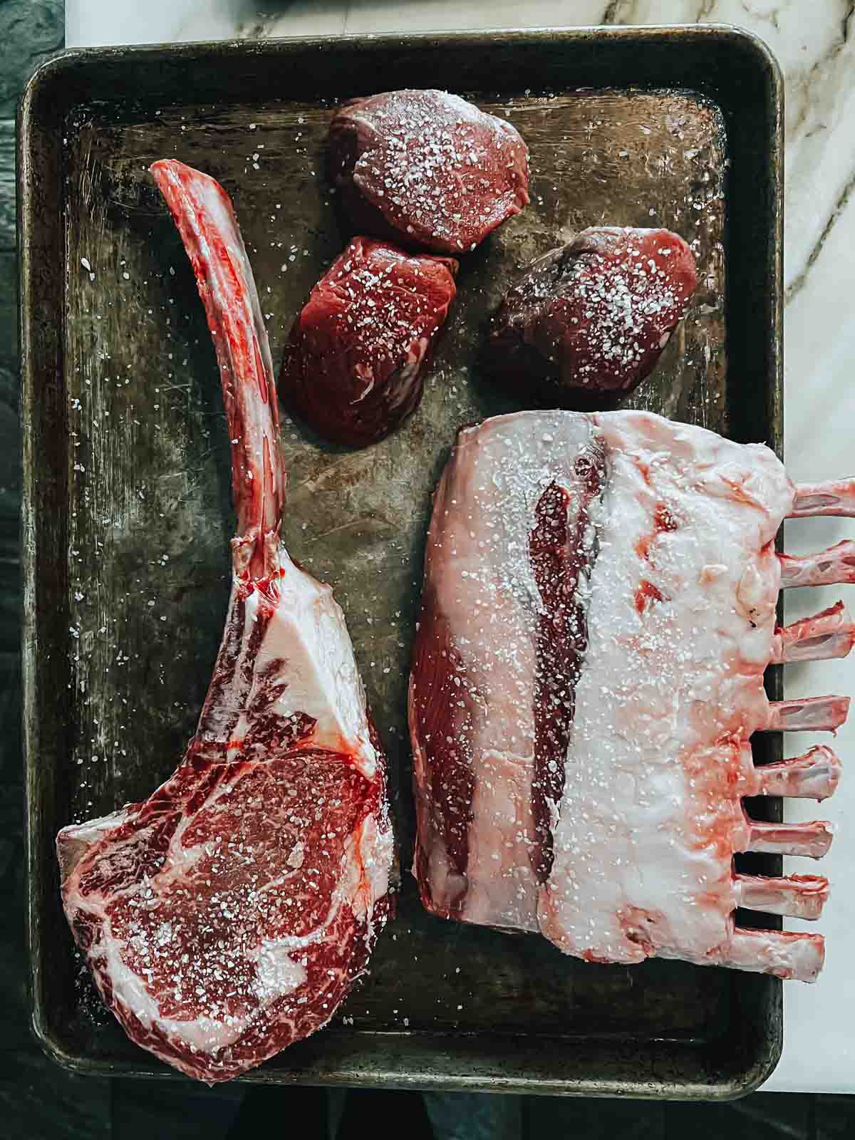 3 cuts of meat on a baking sheet, filets, tomahawk, and rack of lamb. 