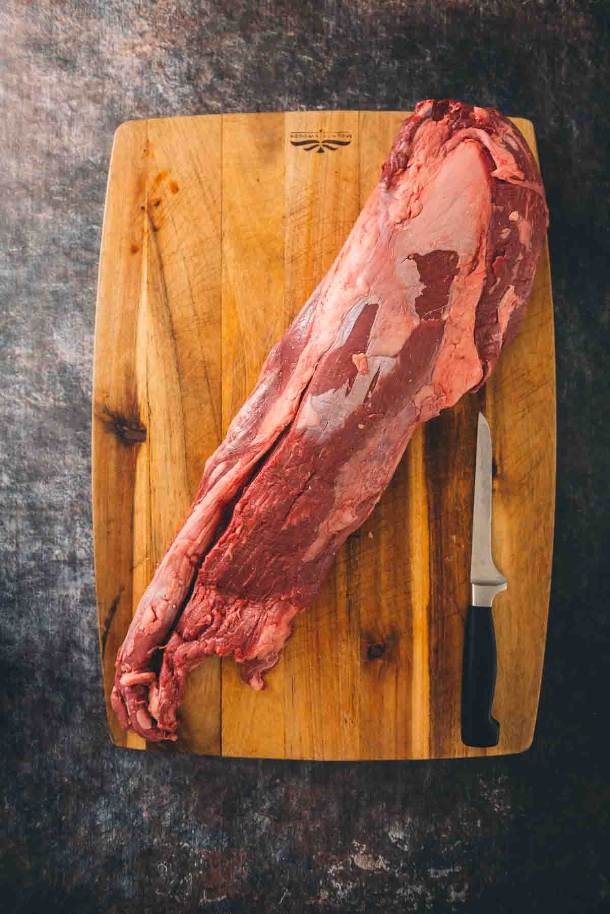 A whole untrimmed beef tenderloin on a cutting board with a filet knife. 