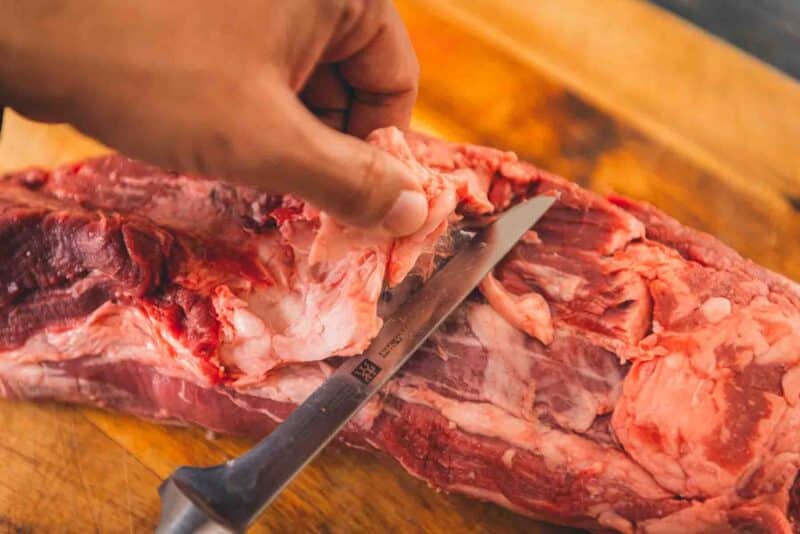 A person trimming a beef tenderloin on a cutting board.