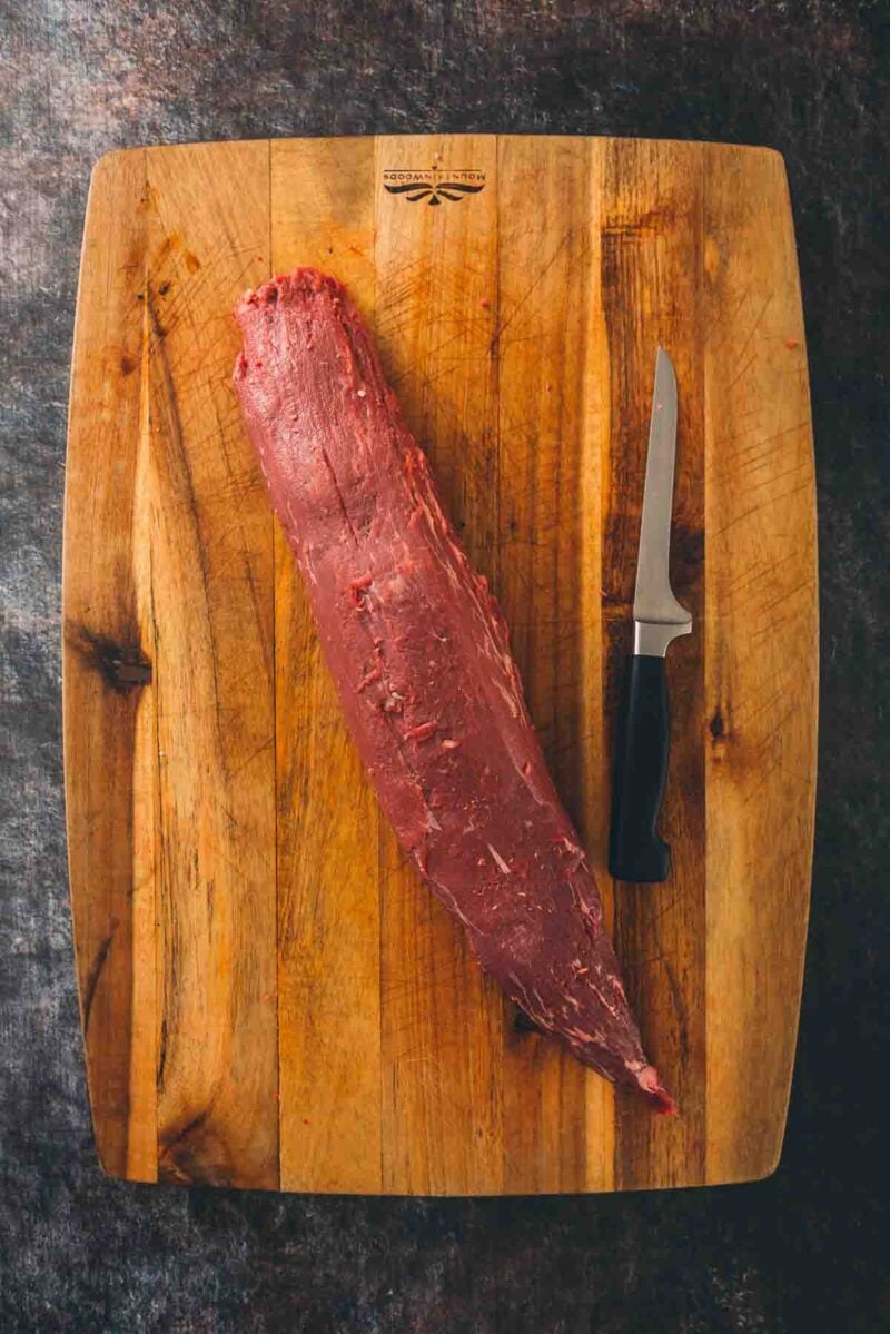 A beef tenderloin being trimmed on a cutting board with a knife.