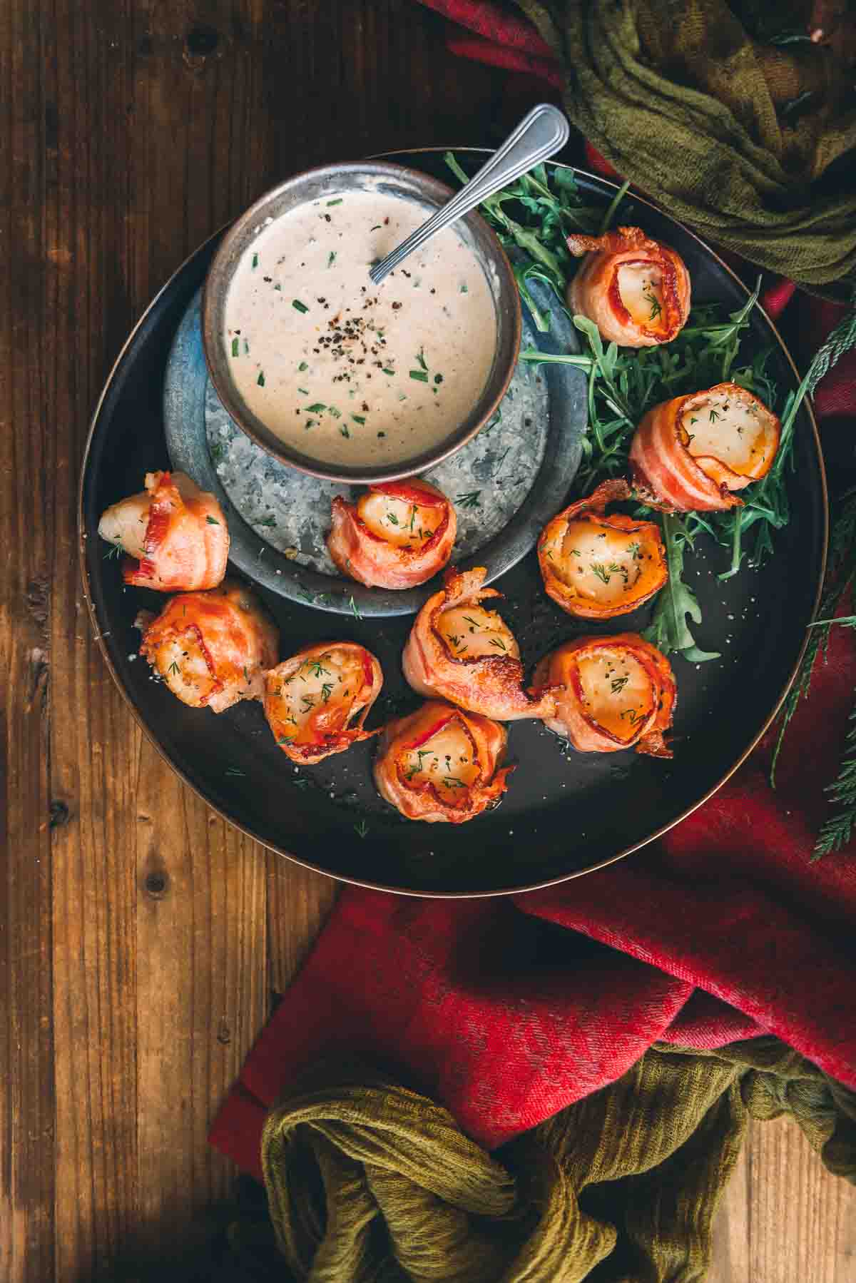 Bacon wrapped scallops on a black plate with a bowl of horseradish dipping sauce for serving. 