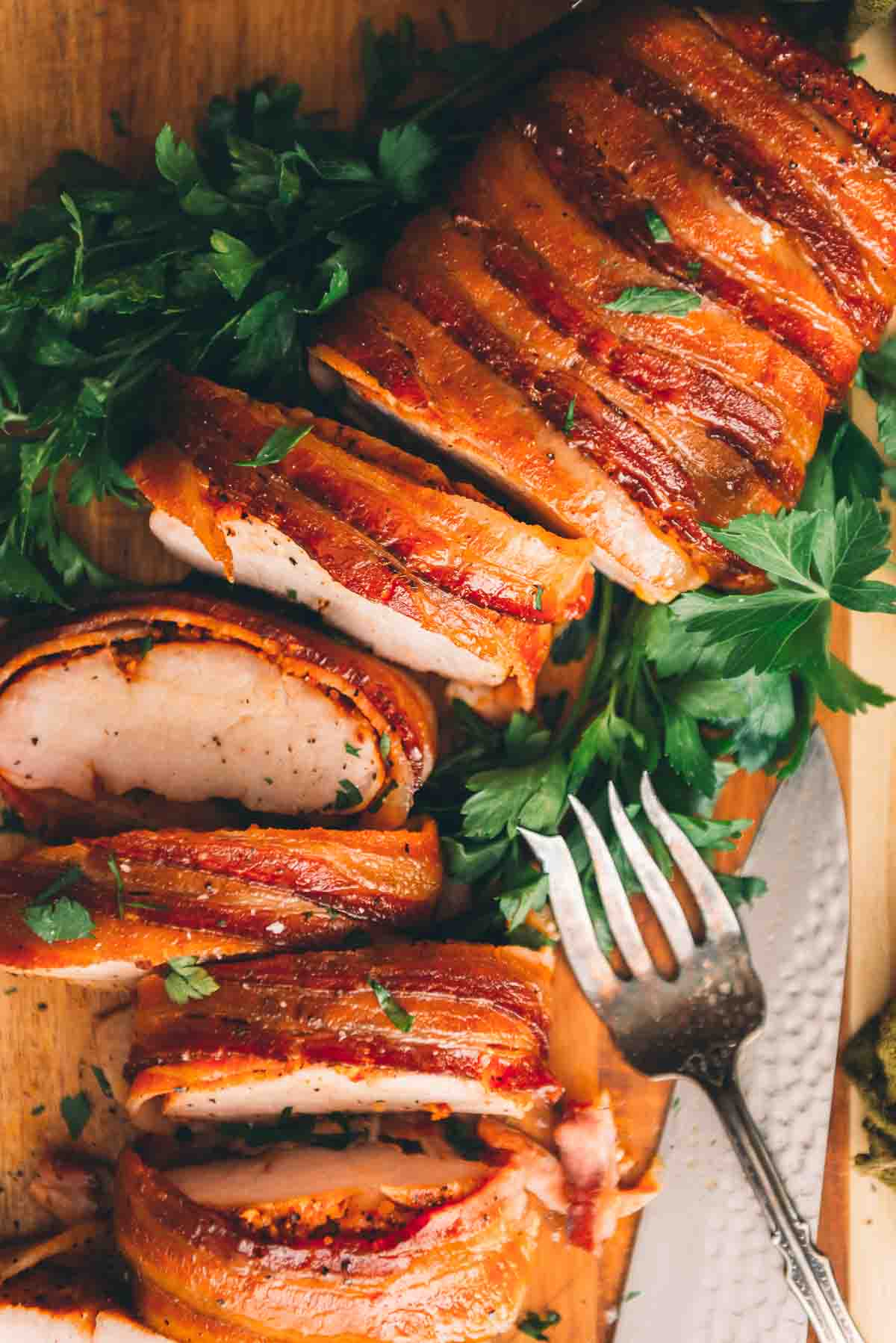Close up of beautifully red smoked bacon layered around a juicy pork loin on a cutting board with parsley. 