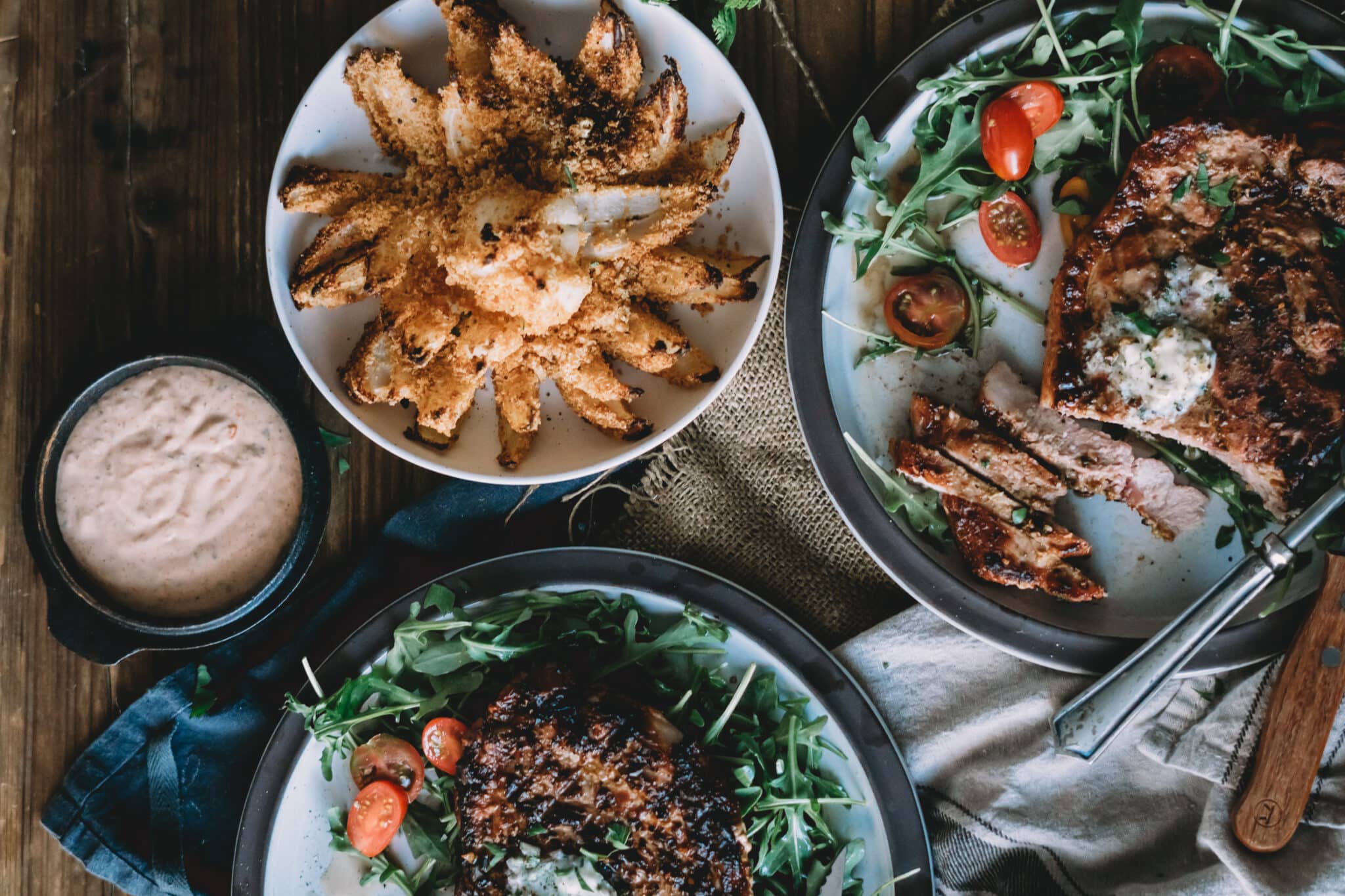 Overhead shot of dinner table with plates of pork chops topped with herb butter, over greens with crispy onion on the side.