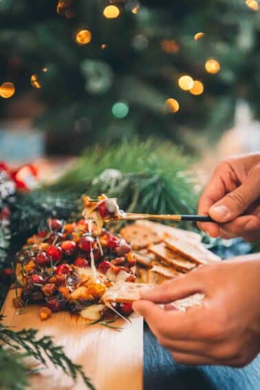 A person is holding a smoked brie pizza on a cutting board in front of a Christmas tree.