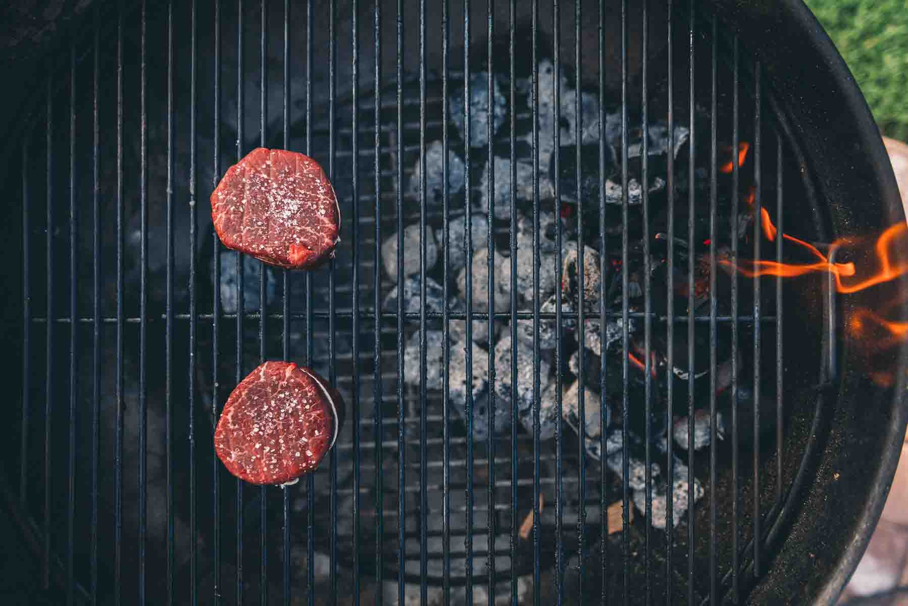 Showing overhead of steaks on the grill, placed on the cooler side, opposite the litcharcoal, of the grill.
