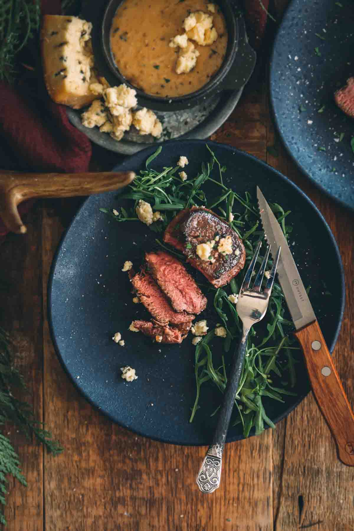 A pan seared sous vide cooked filet mignon sliced to serve on a black plate with baby arugula and gorgonzola. 
