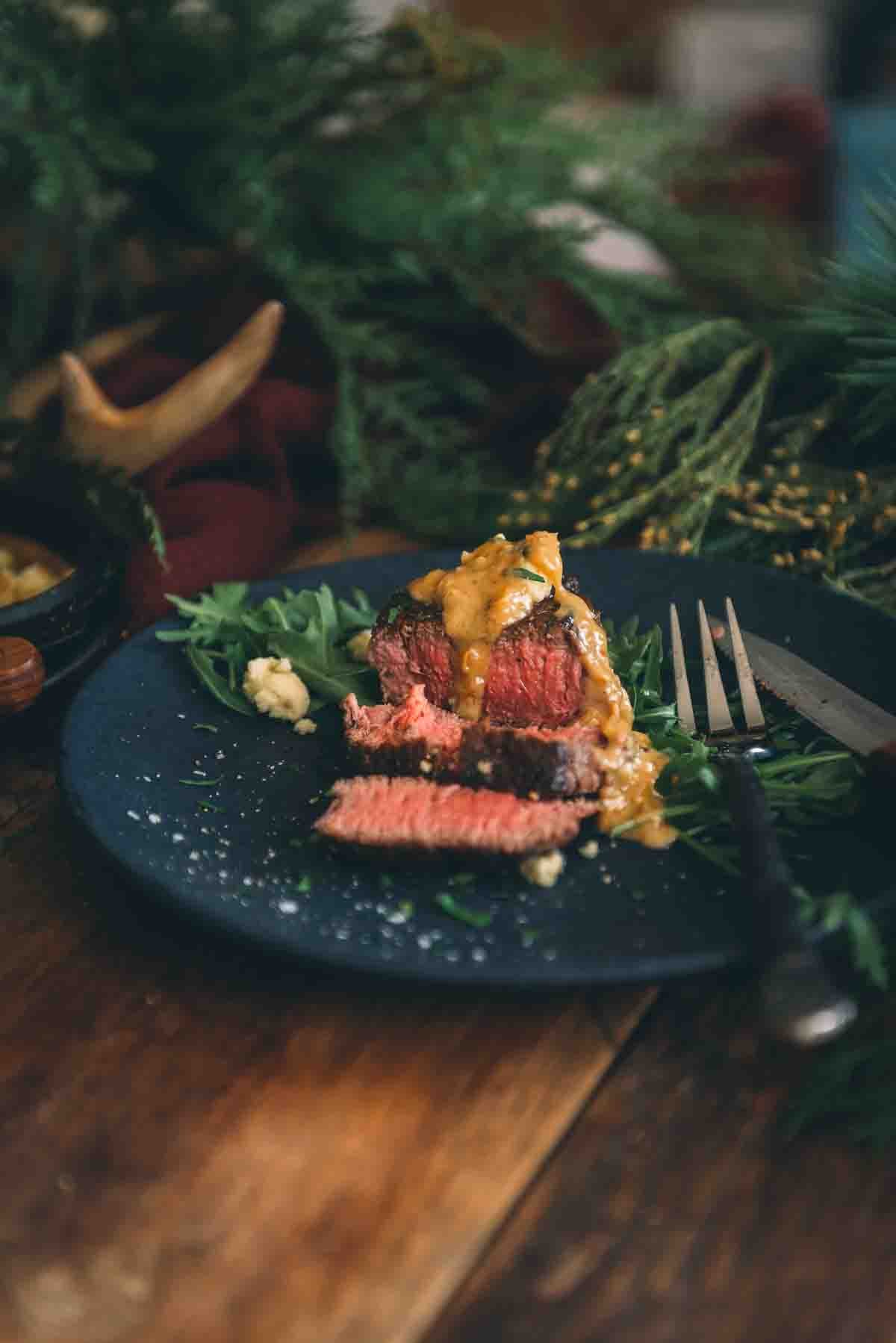 An evenly cooked medium-rare steak on a black plate with blue cheese sauce on top. 