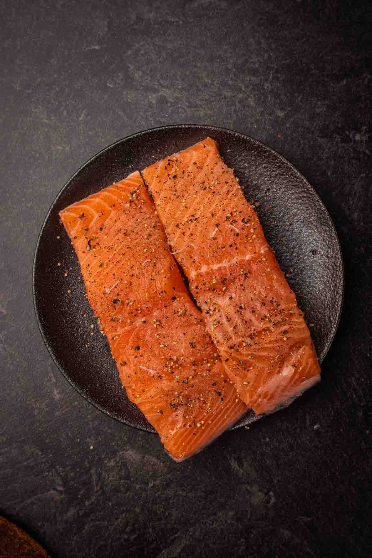 Salmon filets on a black plate, seasoned with salt and pepper. 