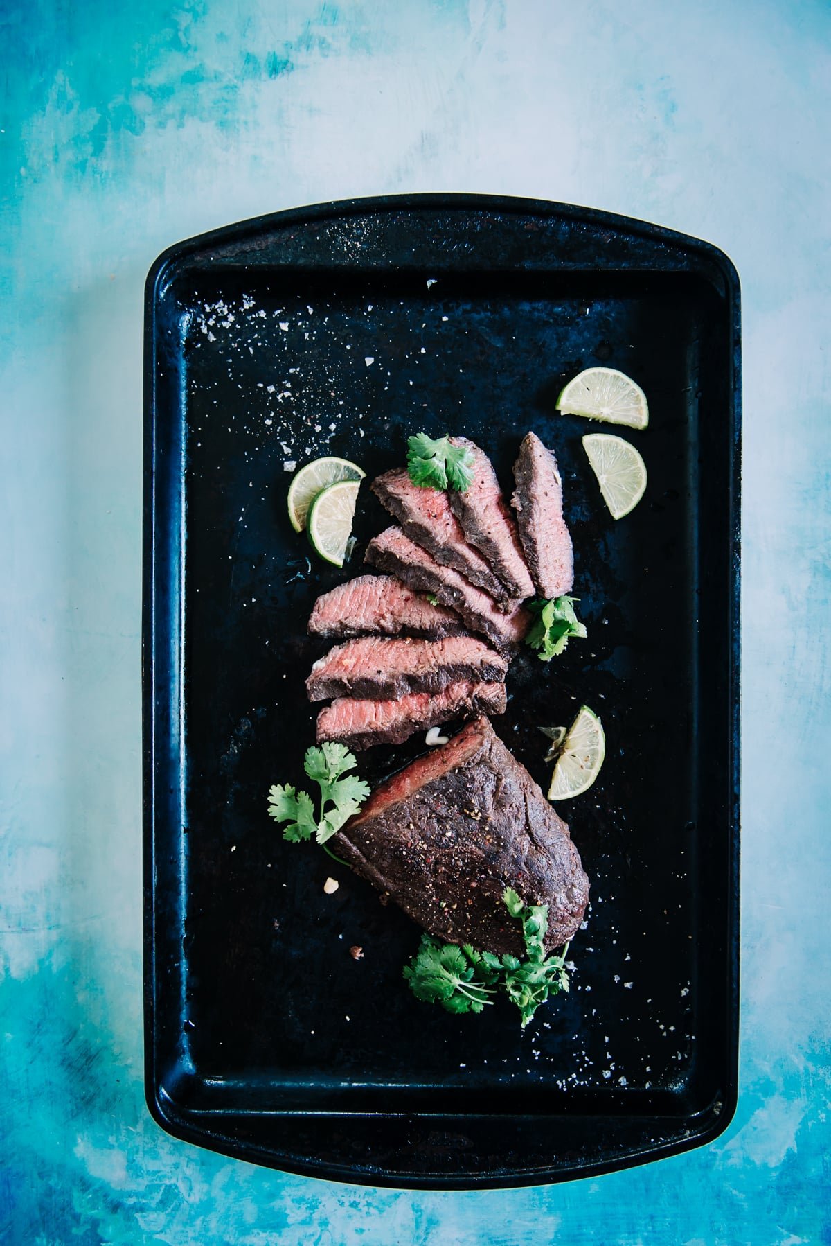 Sliced flat iron steak on a black baking sheet with limes. 
