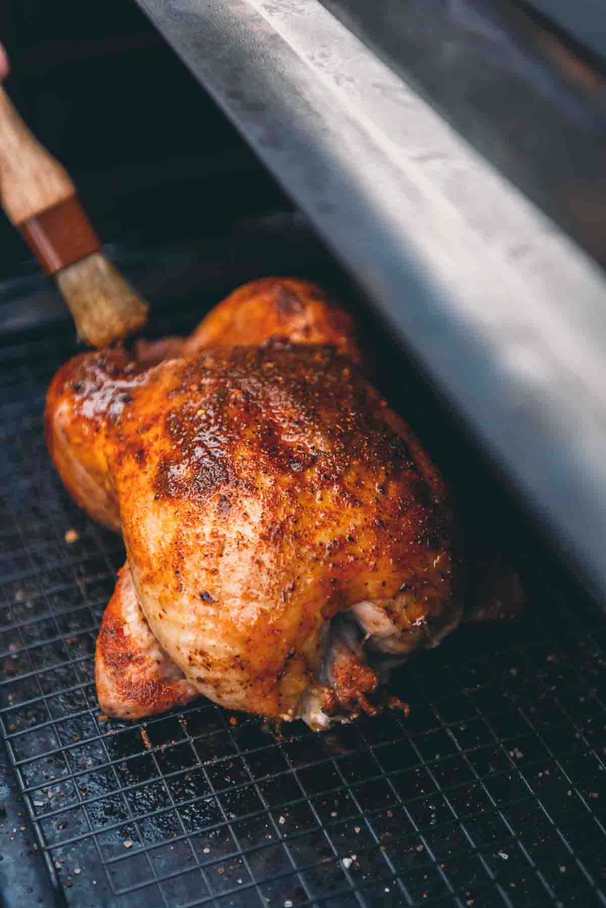 Whole chicken on a grill being basted with a brush.
