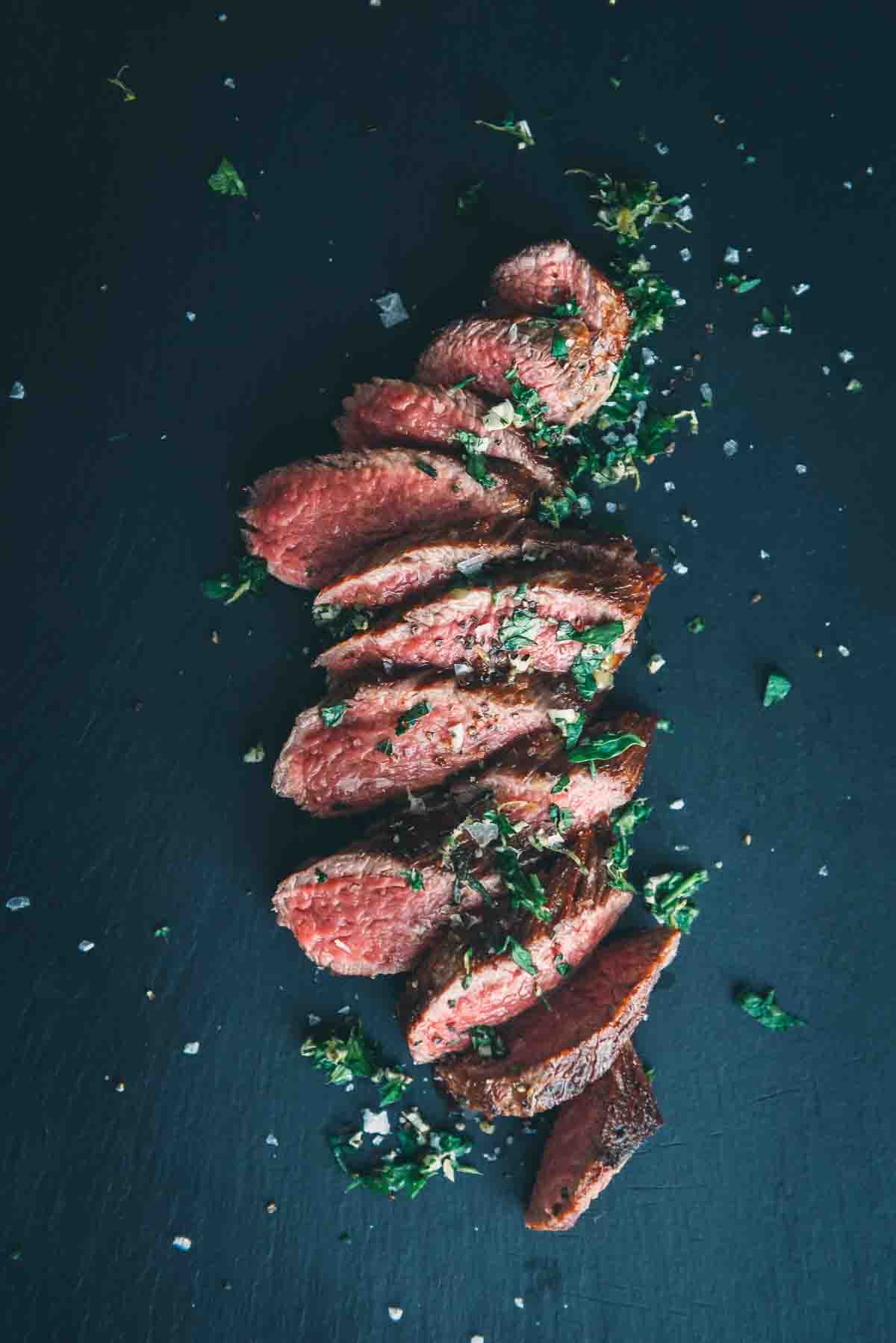 Overhead shot of a cooked teres major steak fanned out on a slate serving platter and garnished with gremolata and salt.
