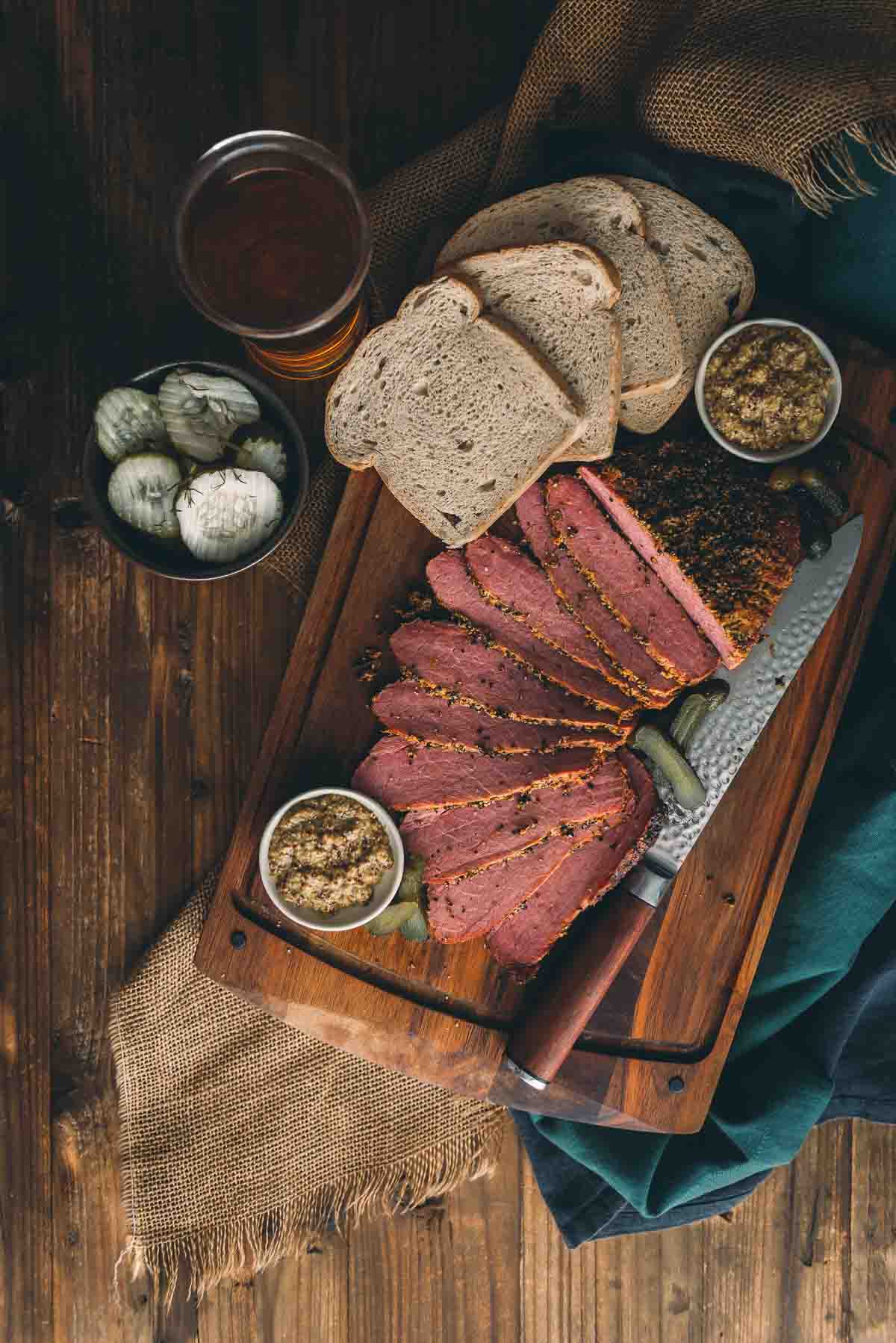 Overhead shot of sliced smoked corned beef on cutting board with bread, mustard and pickles. 