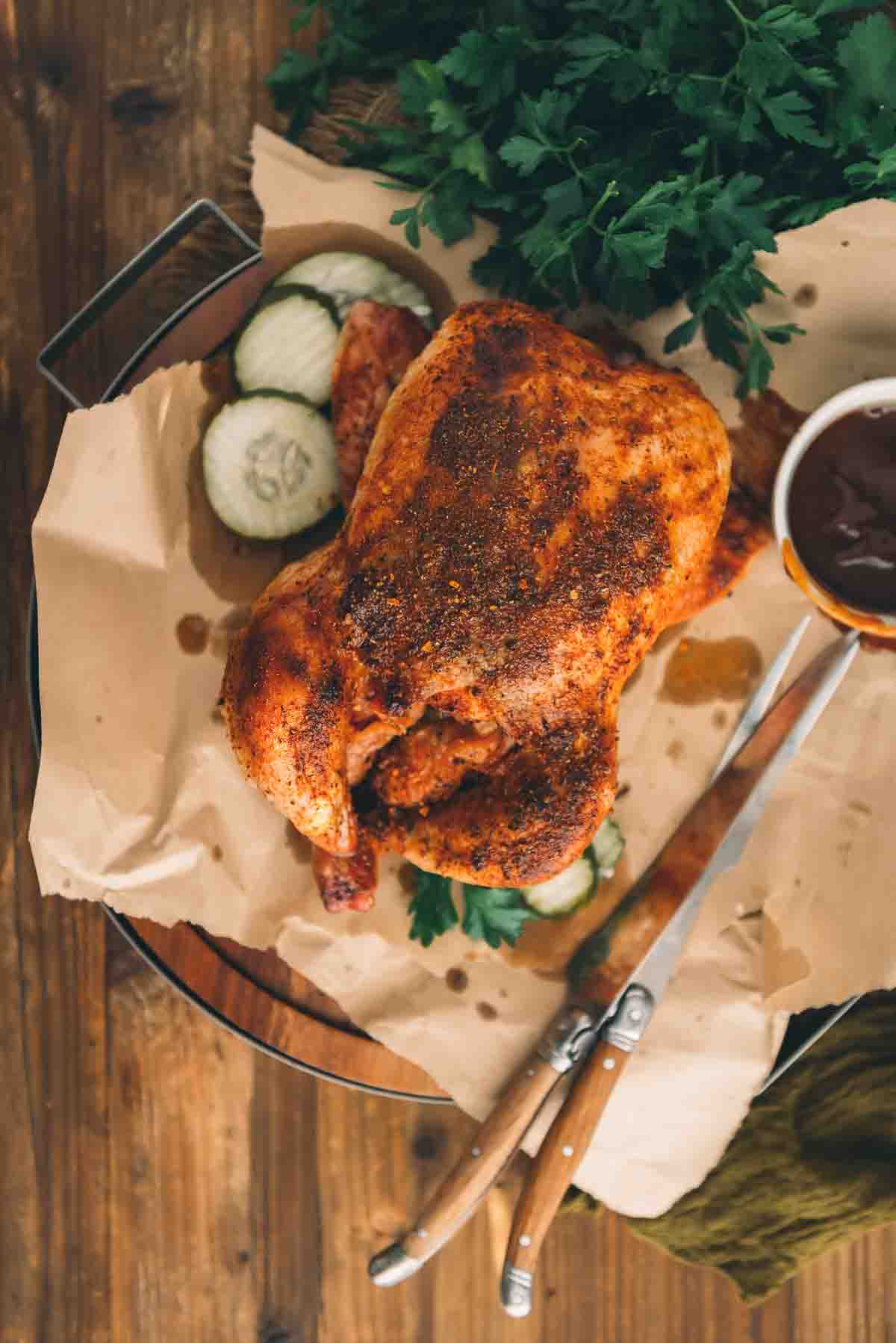 Overhead shot of a golden chicken with spice rubbed crispy skin, arranged on a cutting board with barbecue sauce and pickles ready to carve and serve. 