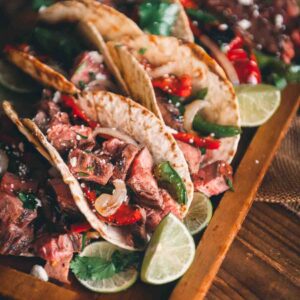 Grilled flank steak tacos in a shell on a platter lined up with lime wedges.