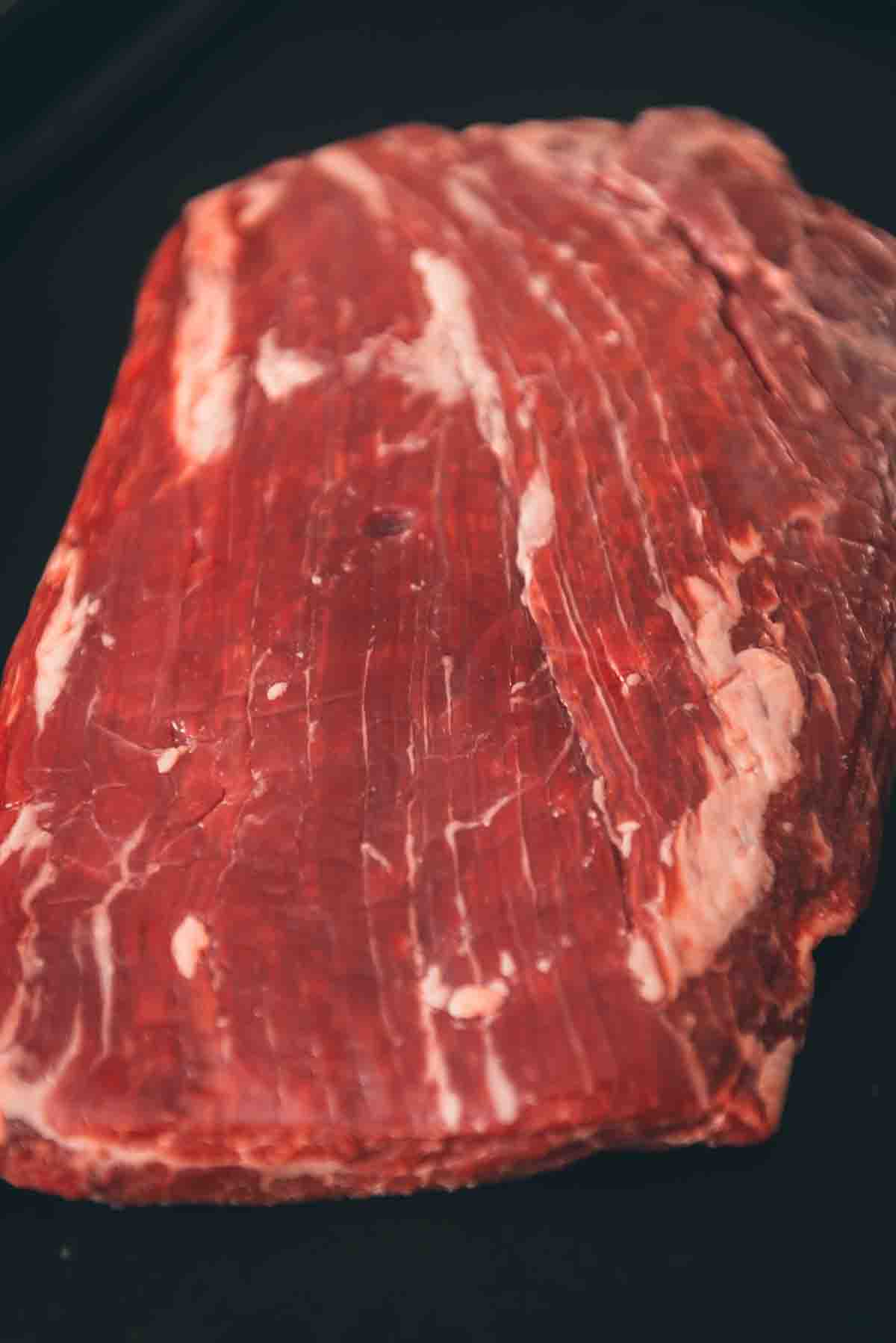 A flank steak is sitting on a black tray to show the long grain. 