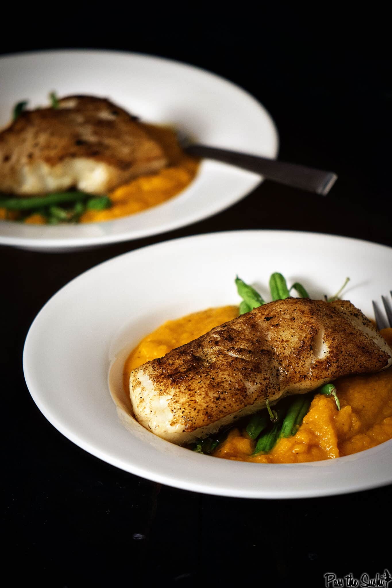 Two white plates with pan seared sea bass fillets served on a bed of mashed butternut squash and green beans, with a fork on the side.