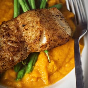 Seared sea bass on butternut mash with green beans, served on a white plate with a fork.