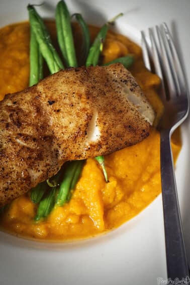 Seared sea bass on butternut mash with green beans, served on a white plate with a fork.