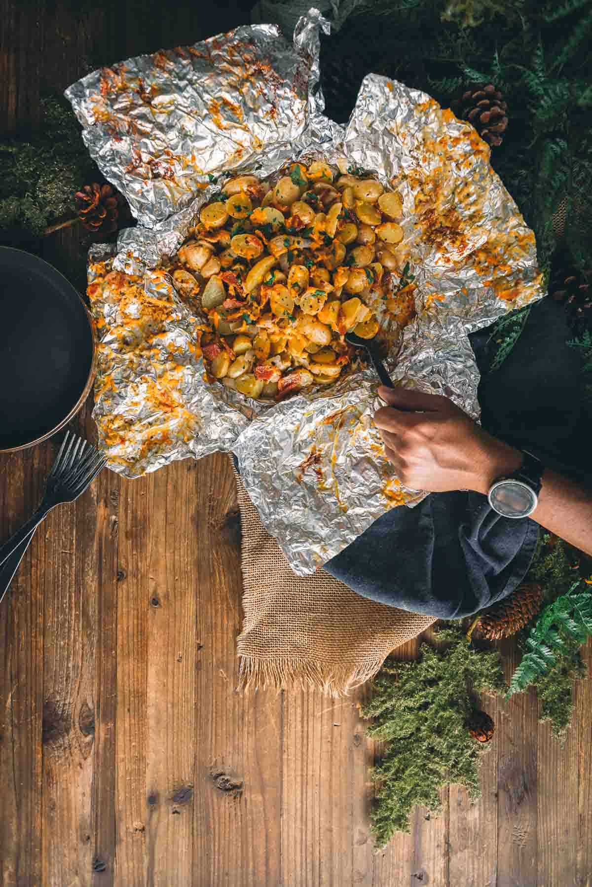 Overhead of open foil packet filled with potatoes with hand scooping some out on spoon.