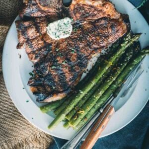 T bone steak with grill marks on a white plate with asparagus and compound butter.