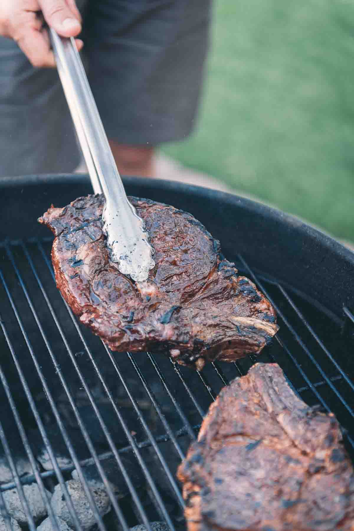 Tongs grabbing a perfectly grilled ribeye steak up off the grill. 