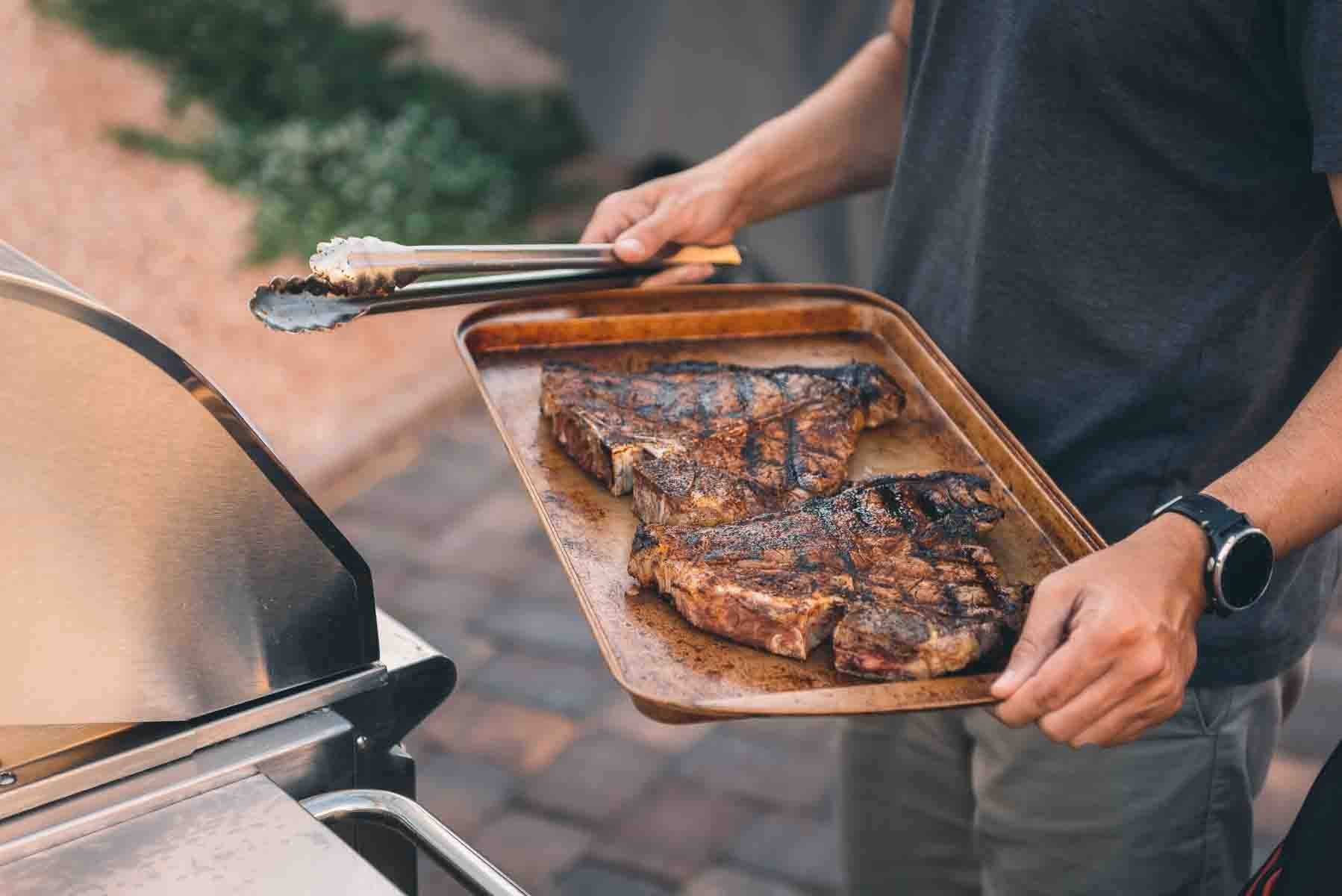 Hands holding tray of grilled t-bone steaks. 