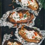 Close up of baked potatoes unwrapped from foil cut open and topped with sour cream, bacon and parsley.