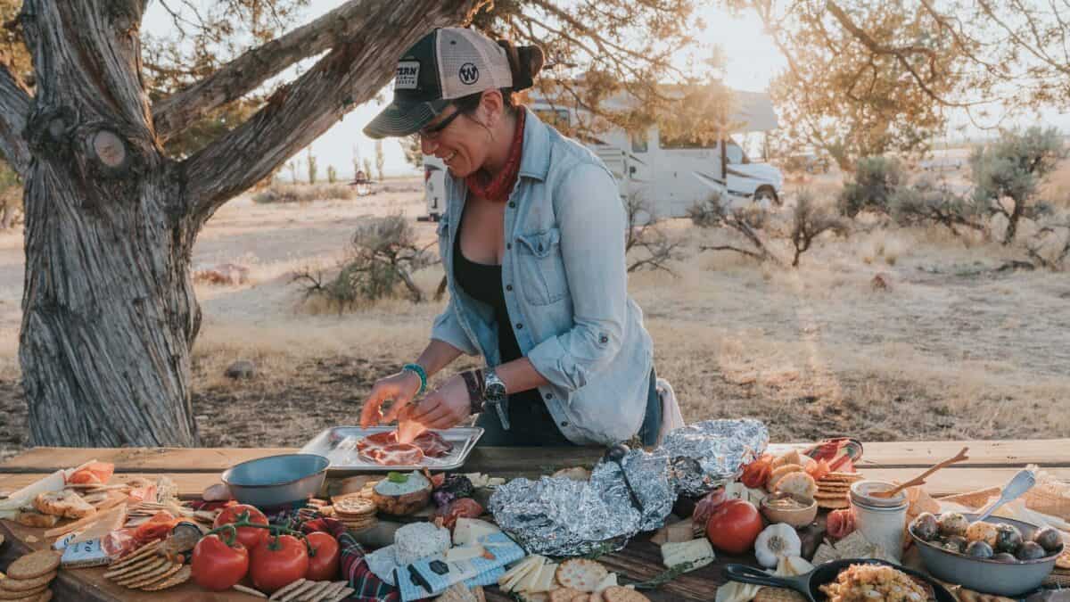 Kita Roberts laying out a large charcuterie spread on a wooden picnic table at golden hour, with an RV and high desert scrub behind her.