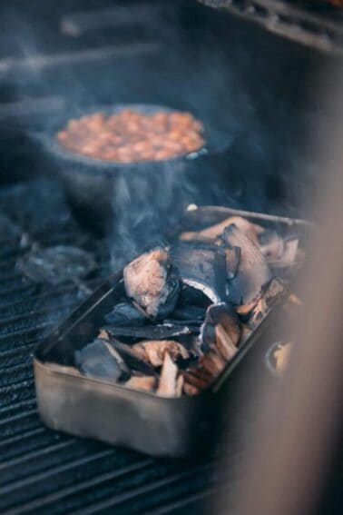 A tin of food on a grill with smoking wood.