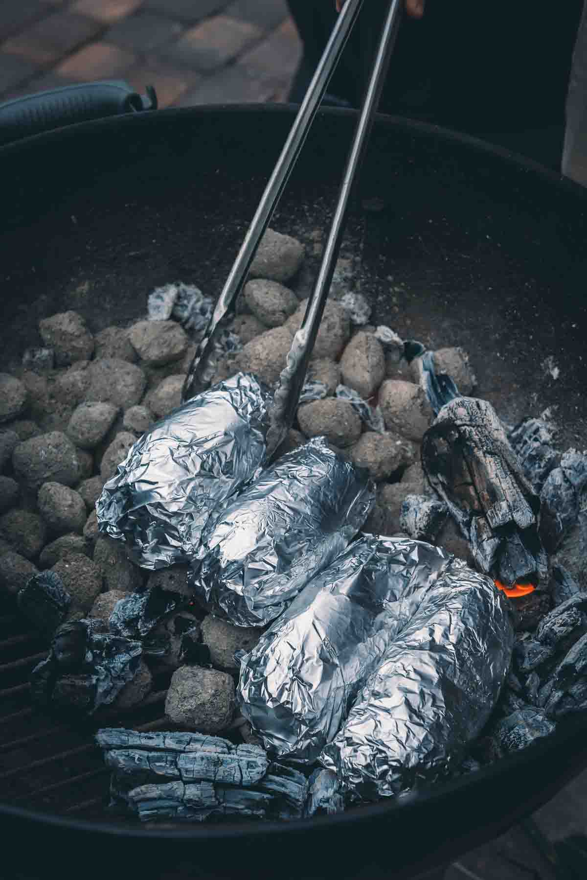 Foil wrapped potatoes being placed in a bed of coals. 
