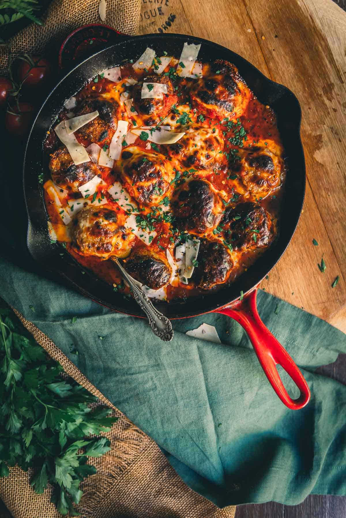 Close up of the skillet of meatballs to show the browned mozzarella cheese and herbs on top in a cozy scene. 