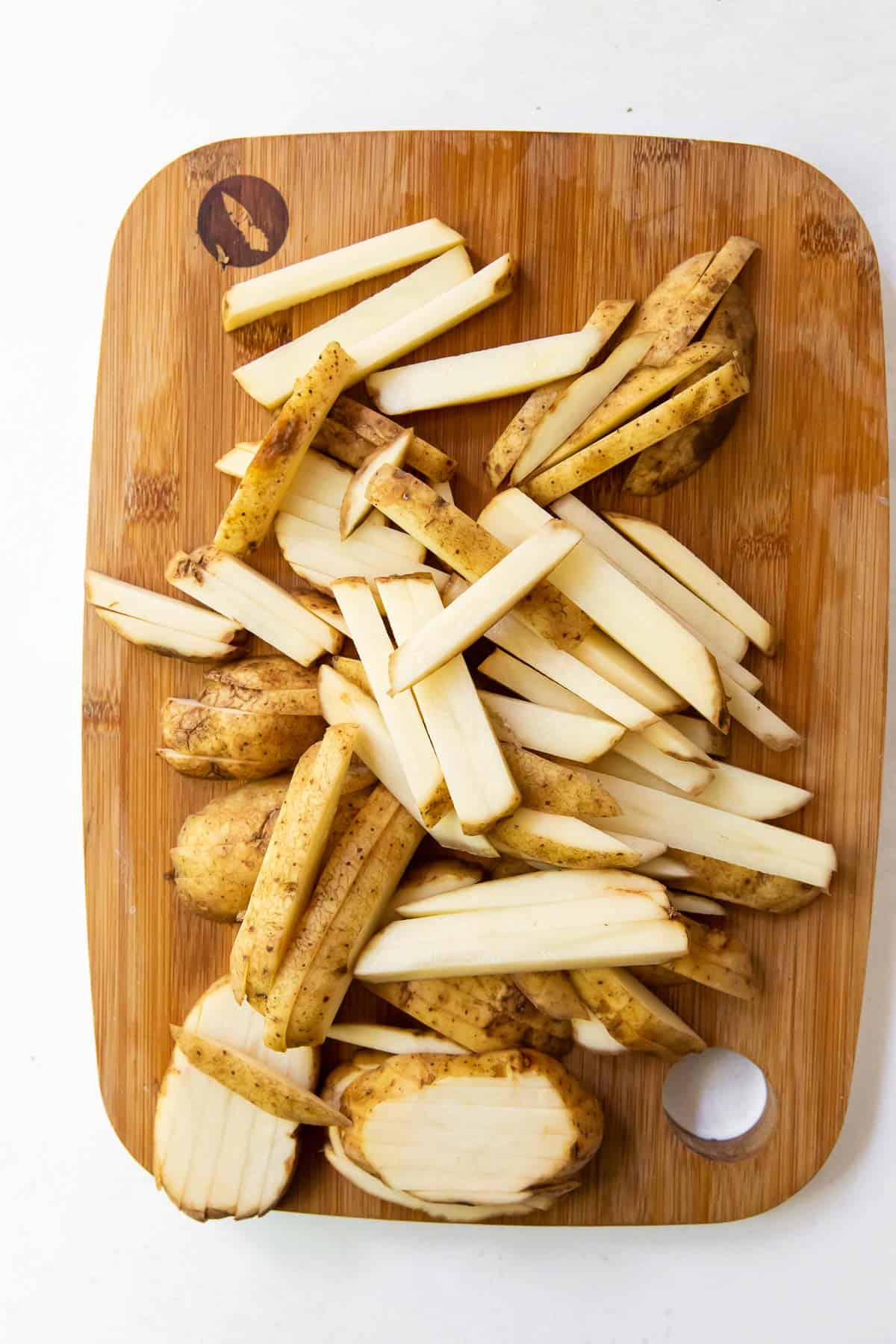 Potatoes sliced into thin fries with skins on on a wooden cutting board. 