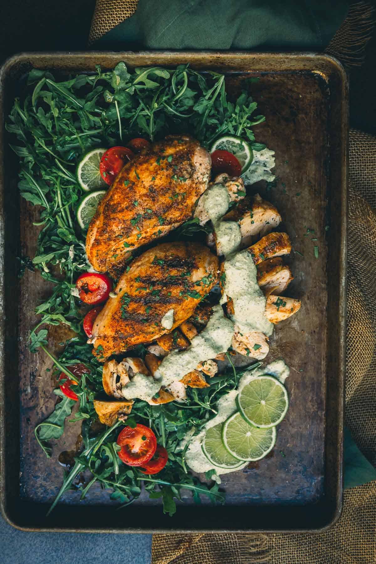 Grilled chicken breasts on a platter with greens, tomatoes, limes and sauce.