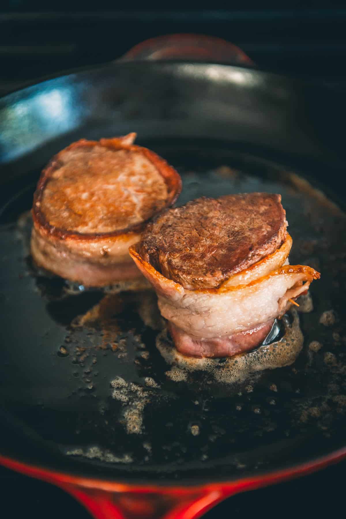 Steaks, wrapped in bacon, being seared in a hot pan.