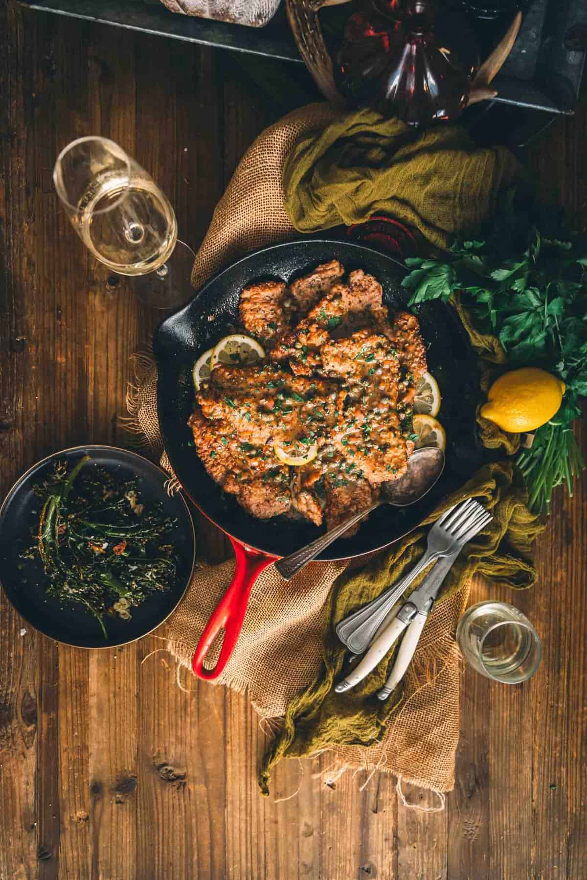 A cast iron skillet with veal cutlets and herbs on a wooden table.