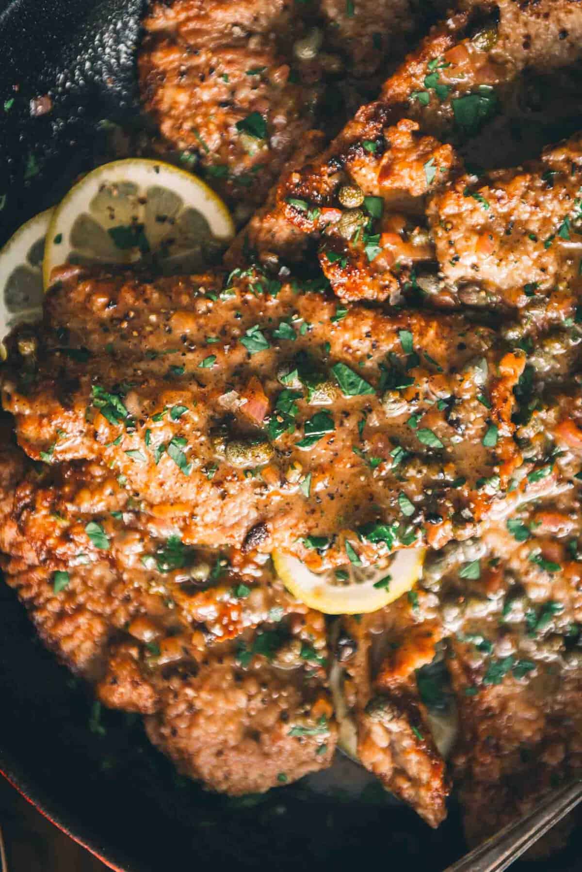 A skillet full of veal with lemon sauce and lemon wedges.