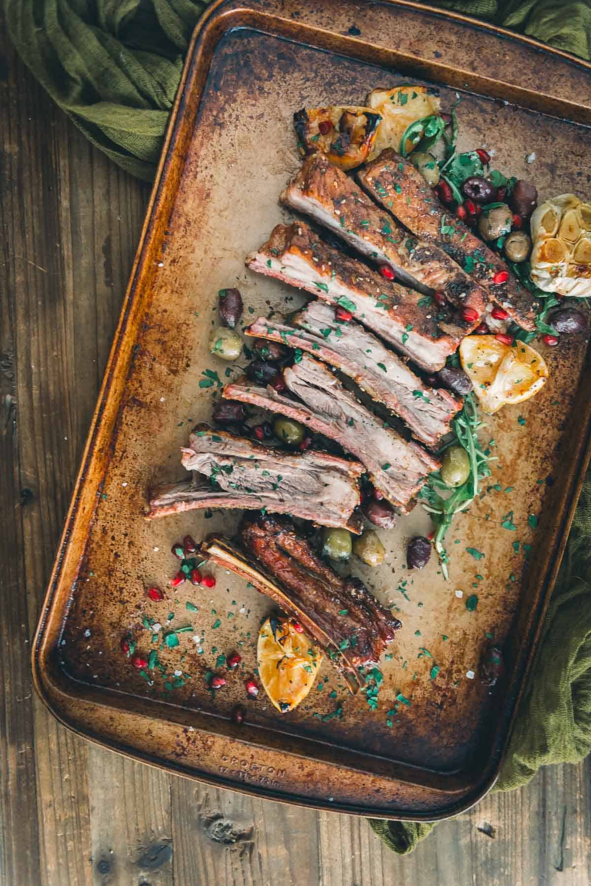 Tender lamb ribs sliced into individual ribs on a platter with olives, garlic and pomegranate seeds.