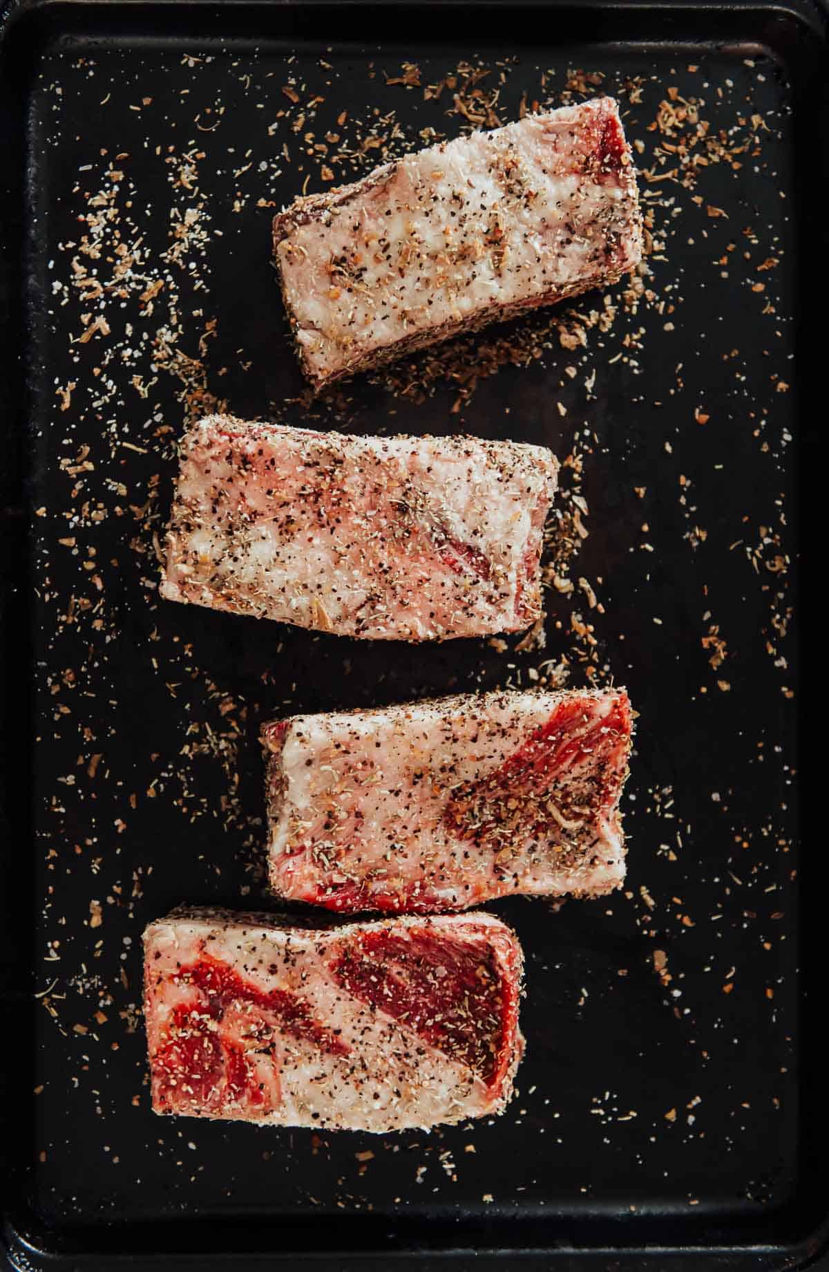 Beef ribs being seasoned on a black tray.