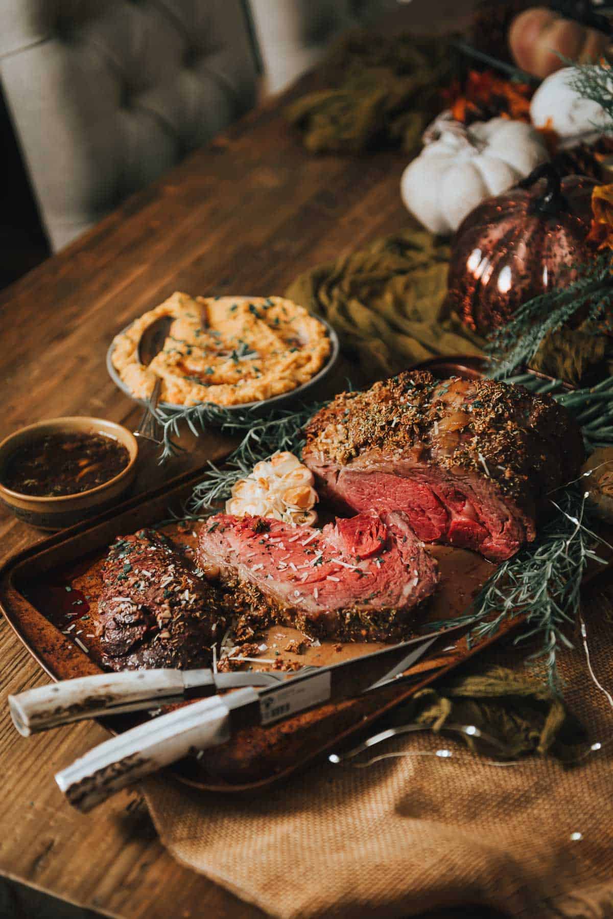 A table with a standing rib roast, vegetables, and a knife.