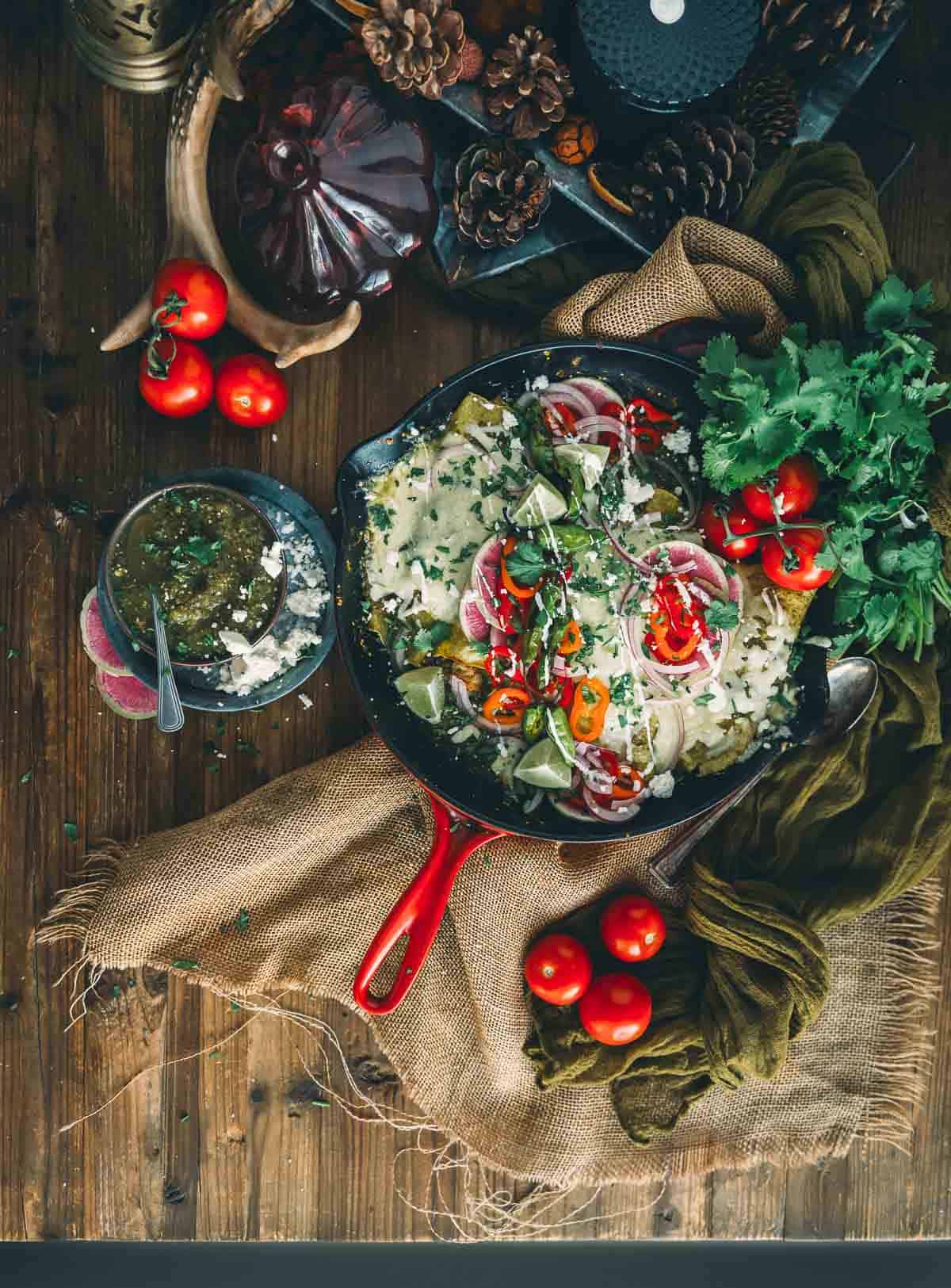 A skillet filled with enchiladas verdes and herbs on a wooden table.