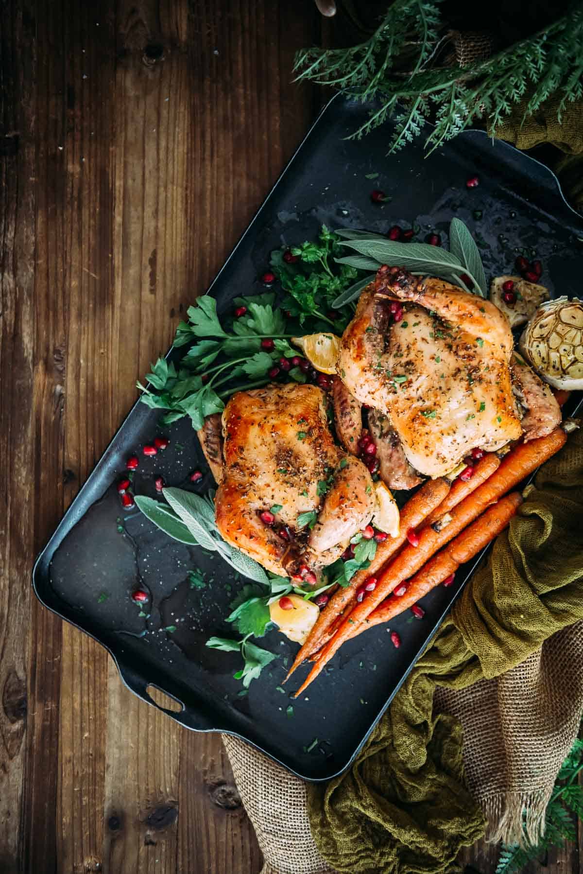 Roasted game hens with carrots and pomegranate on a black tray.
