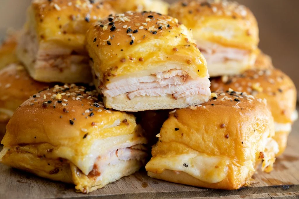 A stack of slider sandwiches with layers of leftover turkey and melted cheese, topped with a sprinkle of sesame seeds and seasonings, arranged on a wooden surface.