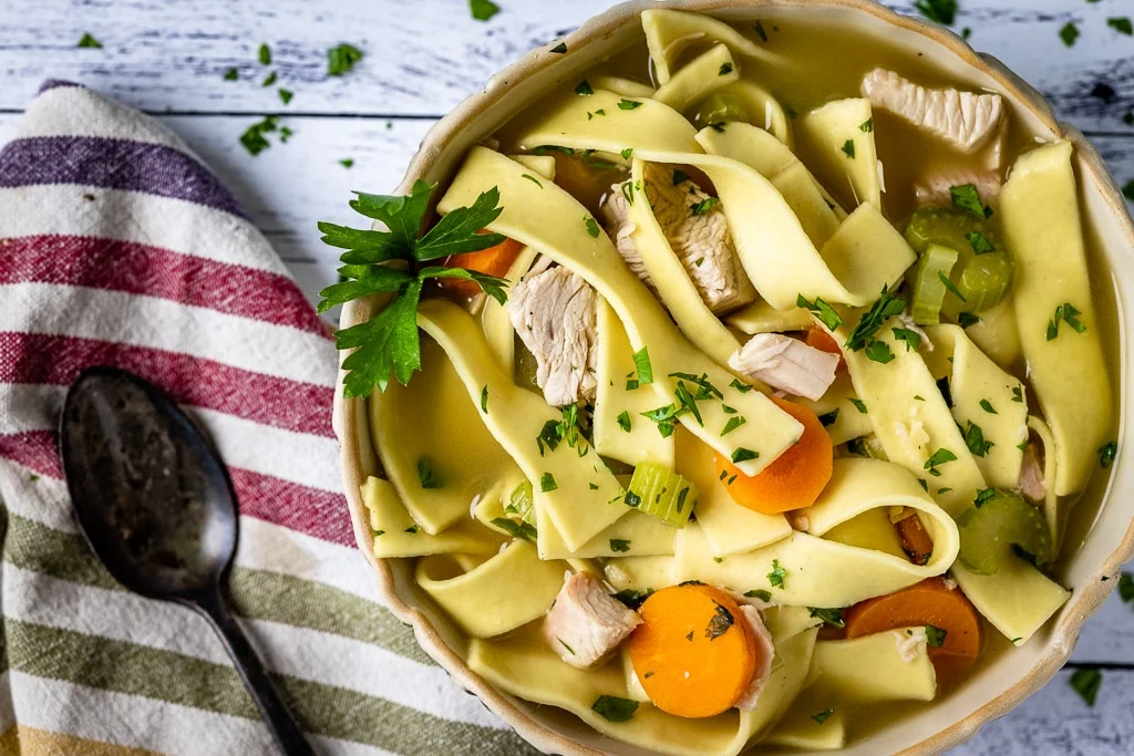 A bowl of chicken noodle soup with sliced carrots, celery, and parsley, placed on a white wooden table next to a striped cloth and a spoon—perfect for those seeking hearty leftover turkey recipes.