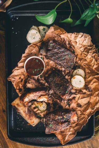 Short ribs on a black tray with dipping sauce on a wooden table.