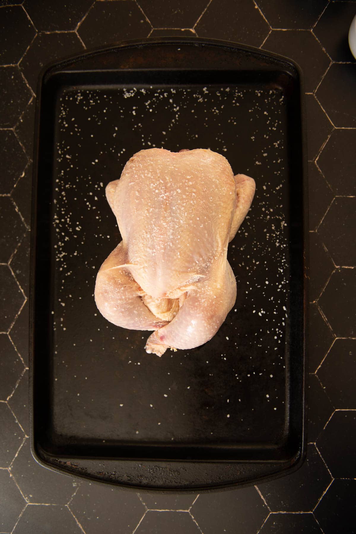 A chicken is sitting on a black baking tray, salted.