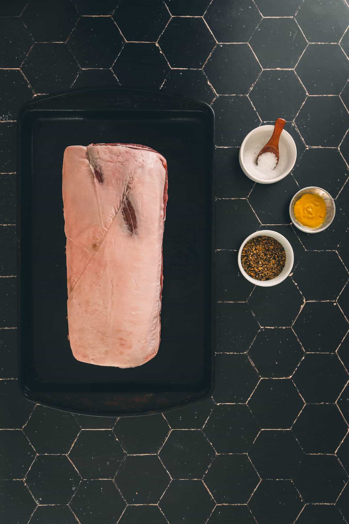 A bone-in pork roast on a tray on a black tiled table.