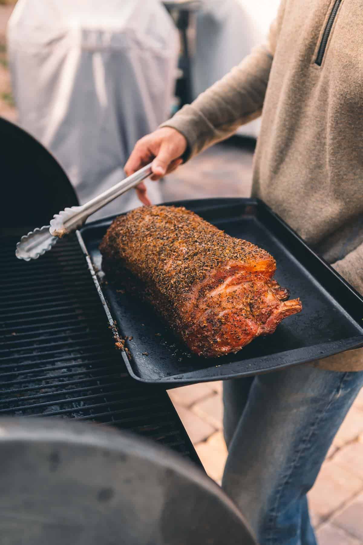 A man is holding taking a rack of pork off the smoker.