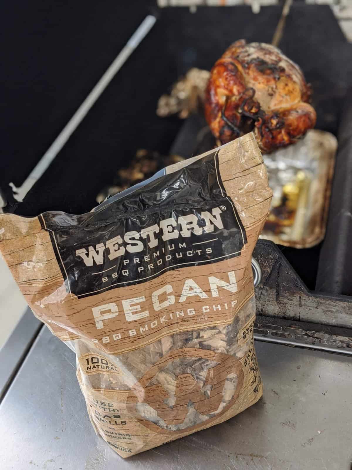 A bag of pecan wood in front of a rotisserie chicken on the grill.