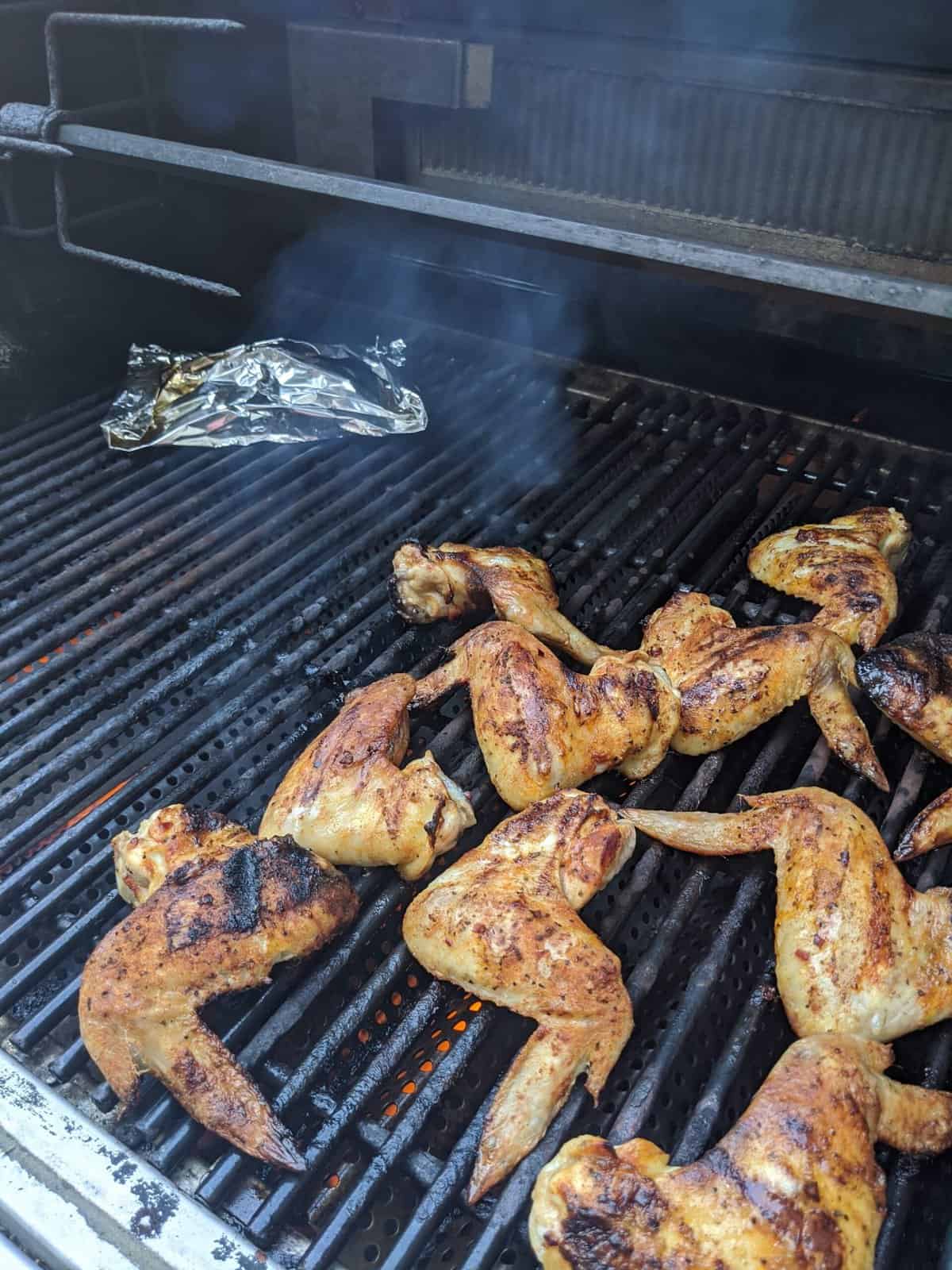 Bbq chicken wings on a grill with a foil packet with the best wood for smoking chicken in the background.