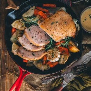 A skillet with pork roast and vegetables on a wooden table.